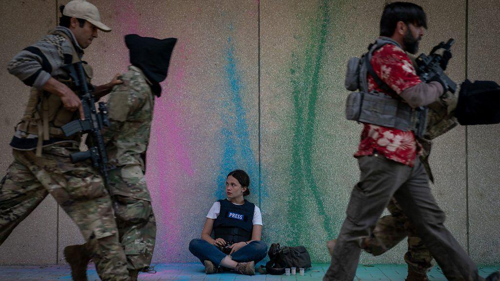 Miltares passam com um preso em frente a uma mulher jovem branca sentada de pernas cruzadas com um colete escrito 'imprensa'