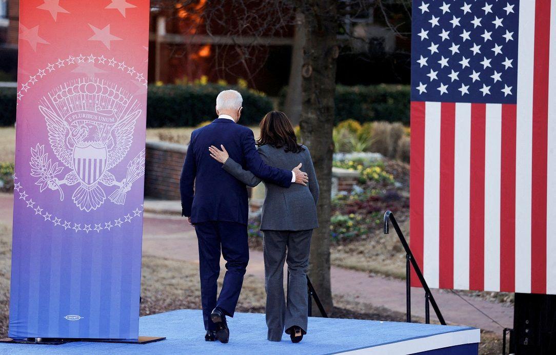 A imagem mostra Biden, um homem branco de cabelos grisalhos vestindo calça e paletó azul marinho, de costas, abraçado com Kamala, que veste calça e terno cinza, saindo de um palco.  Do lado esquerdo, uma bandeira com um emblema, e do outro, a bandeira dos EUA