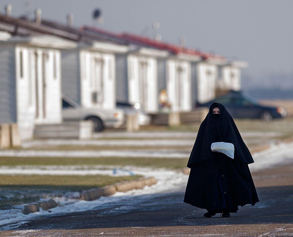 Membro da comunidade Lev Tahor, provavelmente uma mulher, completamente coberta da cabeça aos pés com roupas pretas. Apenas seus olhos e testa são visíveis. Ela caminha por uma rua nevada.