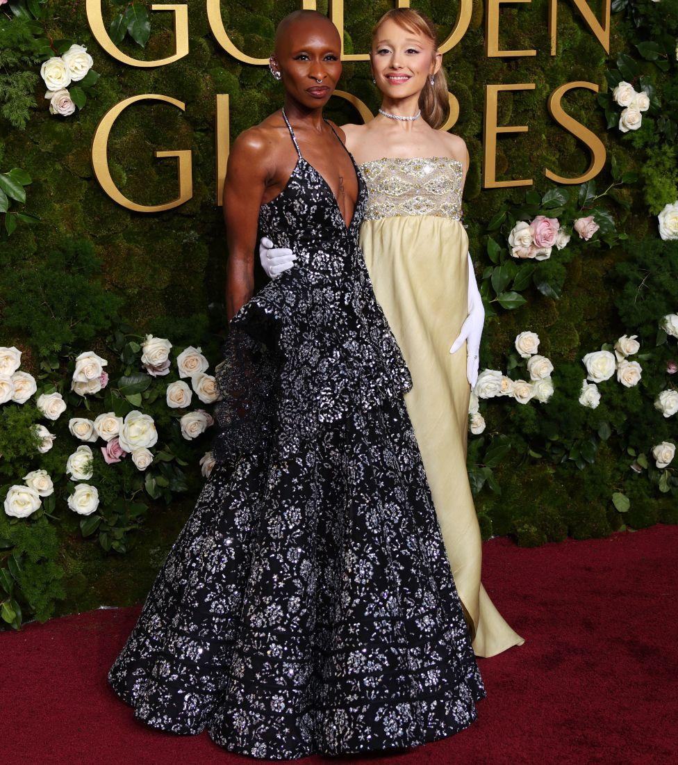 Cynthia Erivo, negra, usa vestido preto estampado com alças, e Ariana Grande, branca, usa vestido claro