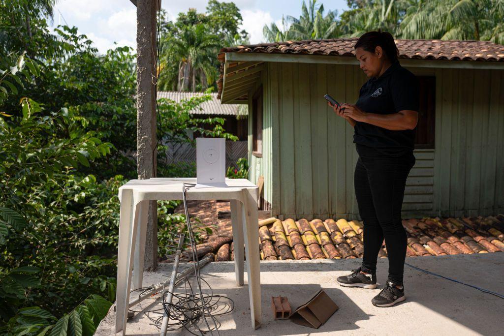 Mulher em pé na varanda, cercada por floresta, em frente a uma mesa de plástico com equipamentos e fios