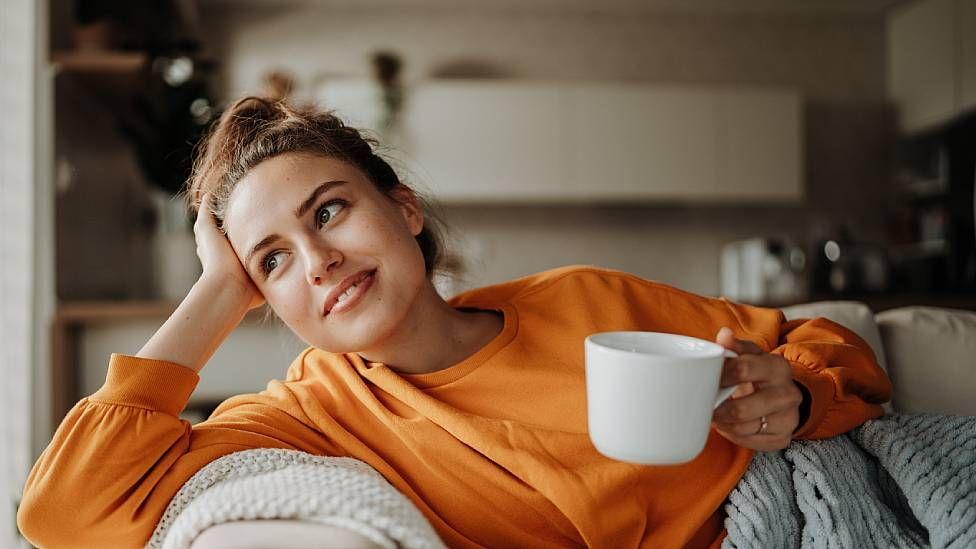 Mujer sonriendo tomando un té sentada en un sillón en su casa