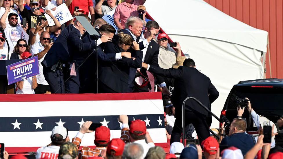 A imagem mostra Trump sendo levado pelos agentes do palco, decorado com as cores da bandeira dos Estados Unidos, rodeado de apoiadores.