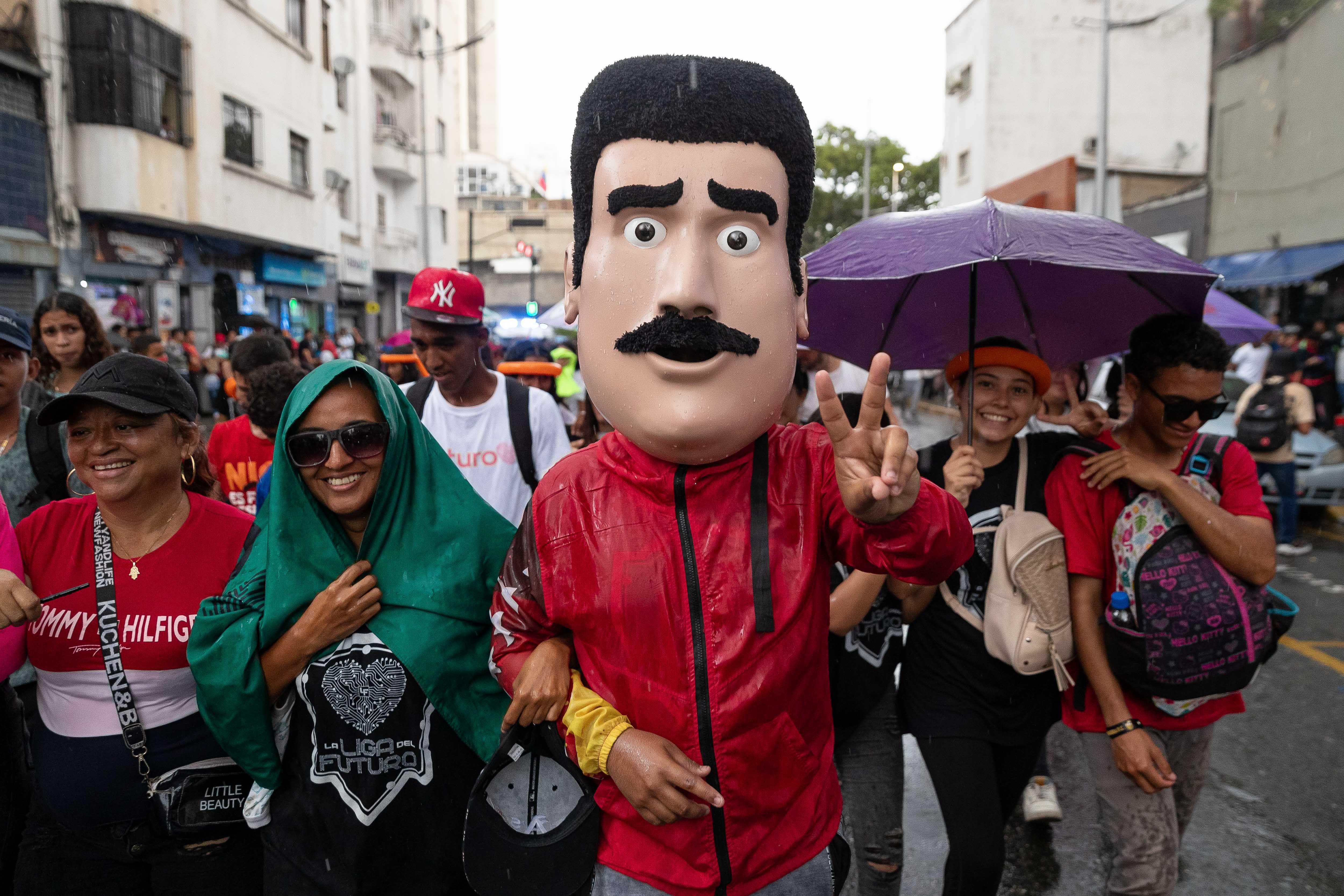 A imagem mostra três pessoas no primeiro plano, uma delas utilizando uma grande máscara de Nicolás Maduro, com bigode e cabelo curto preto.