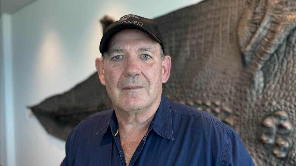 Foto Mick Burns, homem de pele e olhos claros, vestindo camisa azul marinho e boné. 