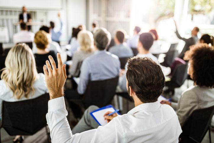 Homem de costas levantando a mão; Com outra mão, ele faz anotação. Ele está em uma sala de reuniões com outras pessoas sentadas e outra posição, liderando a reunião