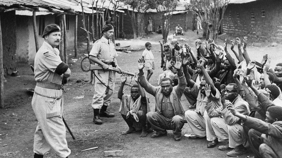 Soldados britânicos em pé diante de rebeldes quenianos sentados