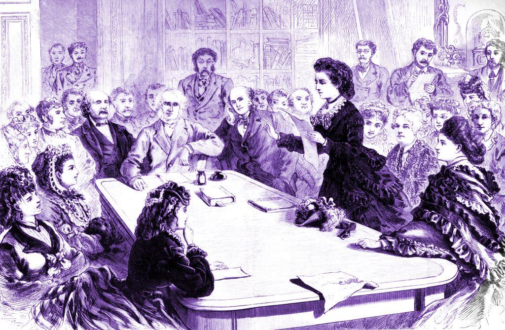 Alrededor de una mesa, hombres y mujeres escuchando a Woodhull, que está de pie
