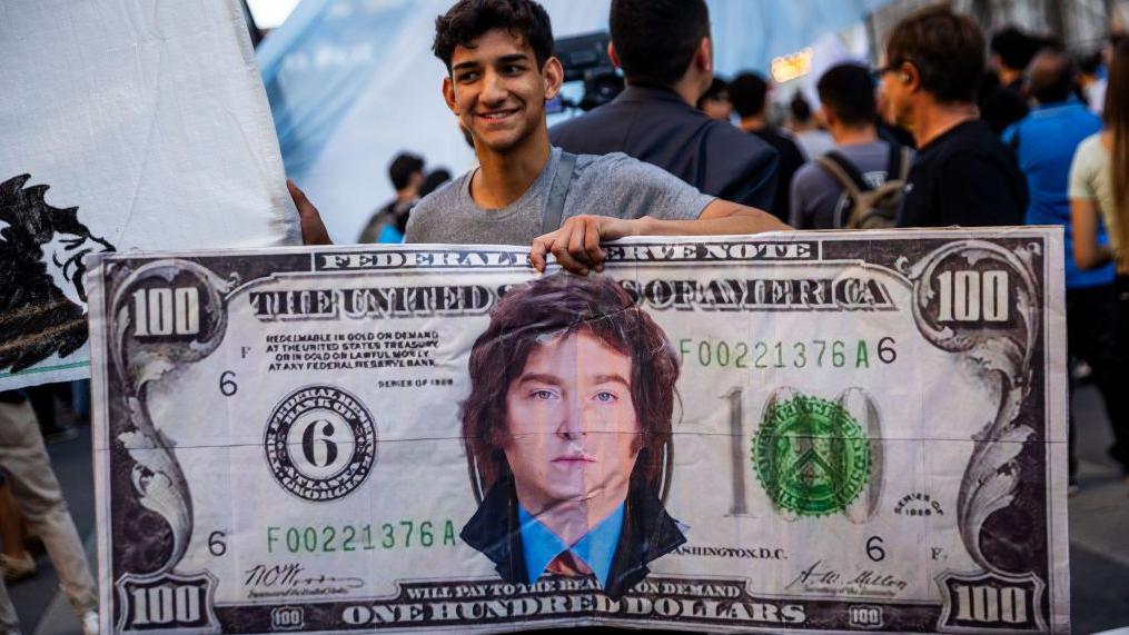 Un chico sostiene un cartel que es un billete de 100 dólares con la imagen de Milei