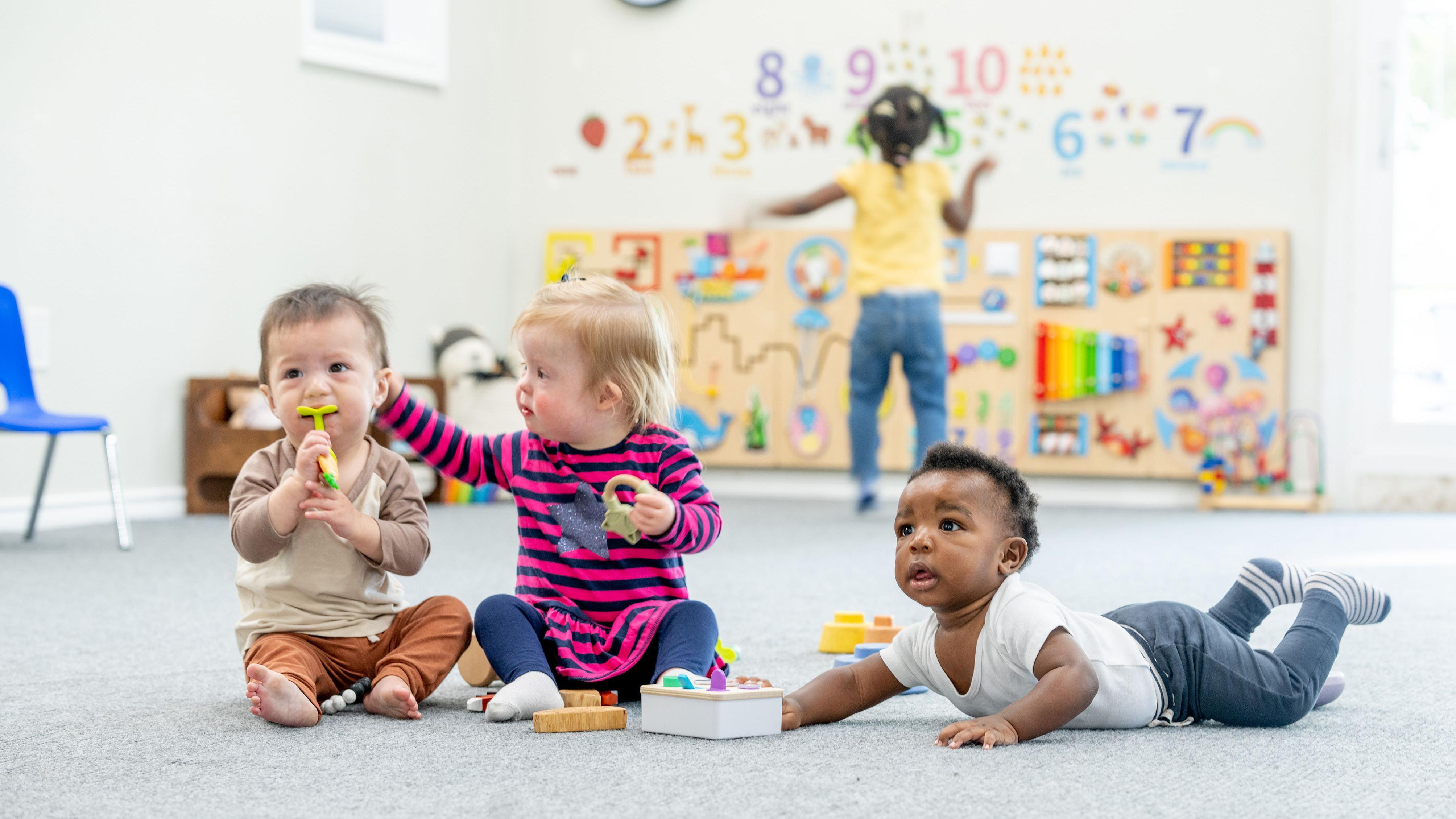 A small group of toddlers sit on the floor of a daycare room as they play together with various toys. 