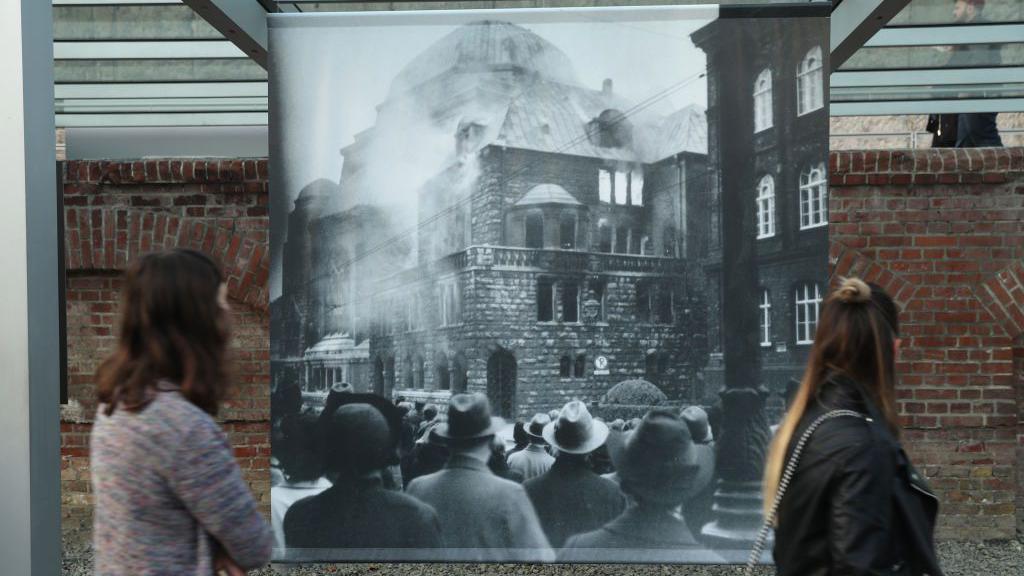 Duas mulheres em frente a grande foto de sinagoga incendiada  