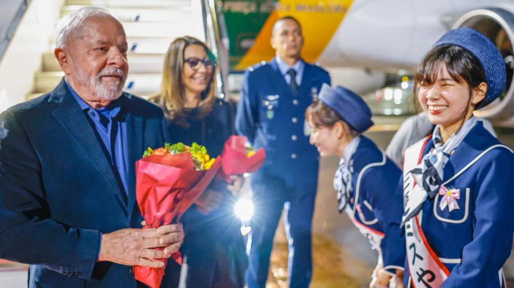 Lula e Janja sorrindo, segurando flores, perto de avião presidencial e de mulheres japonesas os recebendo