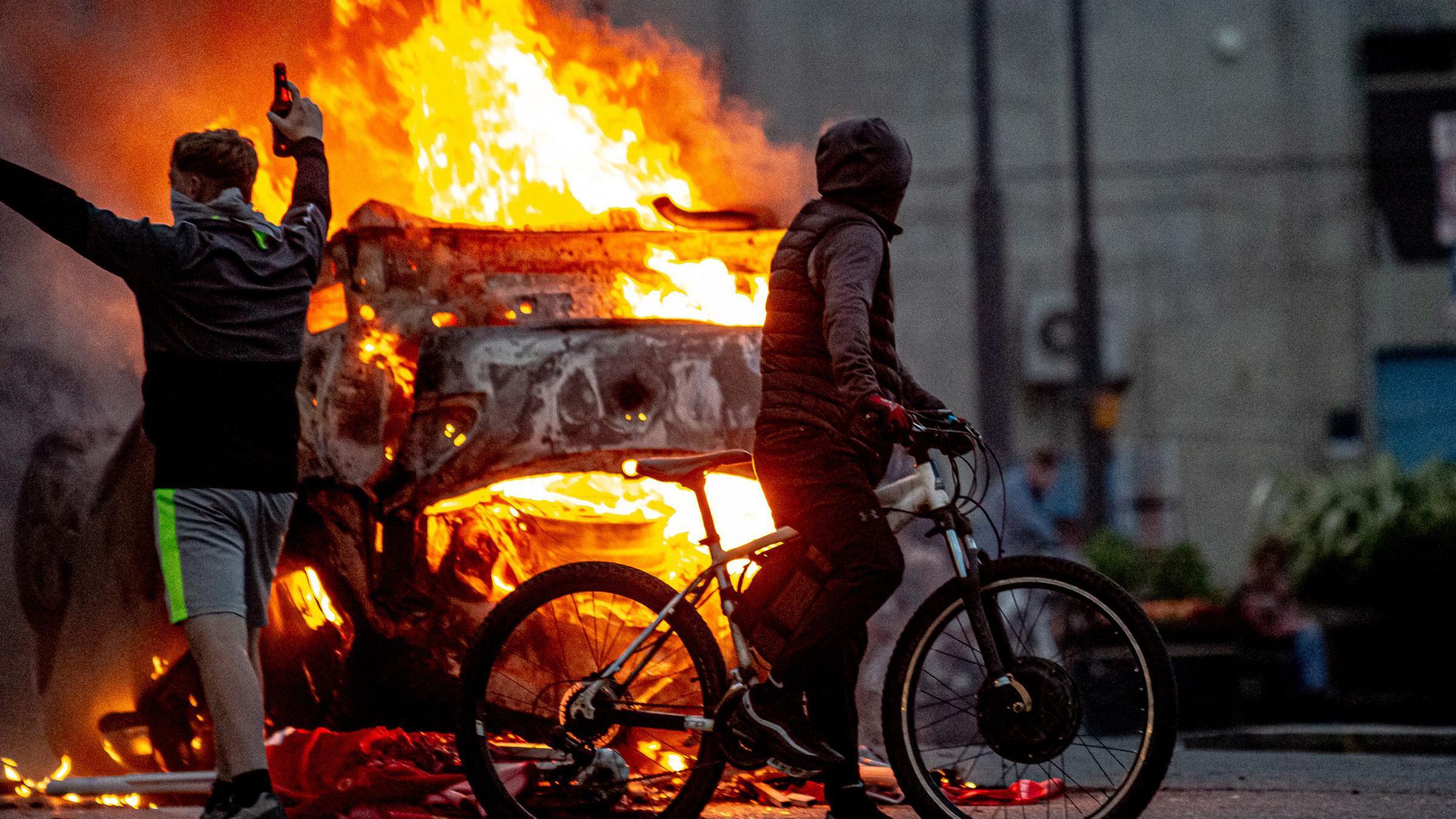 Hooded youths including one on a bike stand next to a burning police car in Sunderland on 2 August 2024