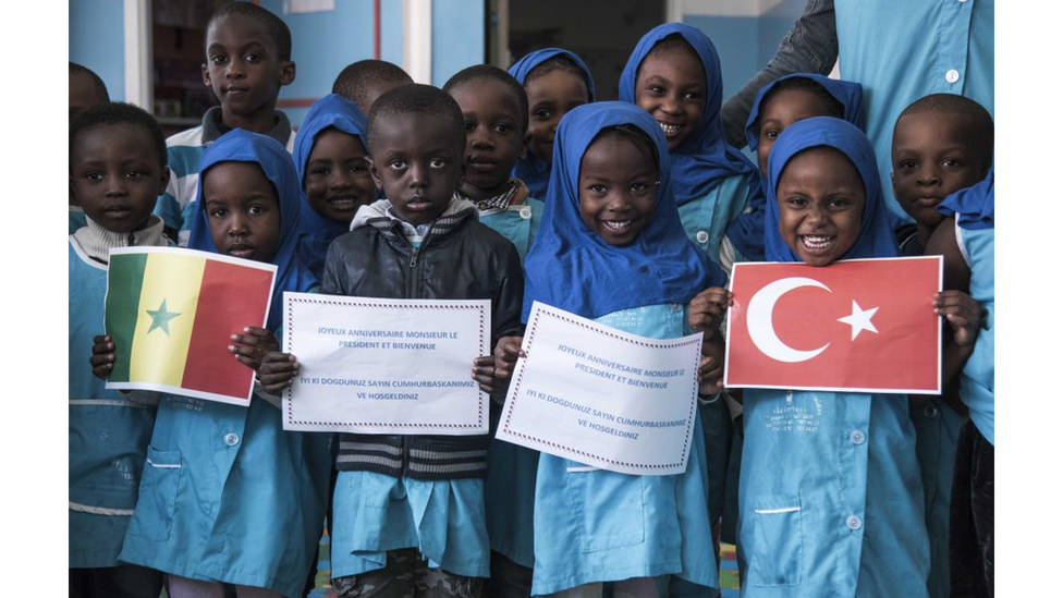 Crianças de uniforme azul em uma escola no Senegal recebendo com cartazes e bandeiras o presidente Erdogan durante sua visita ao país em 2018