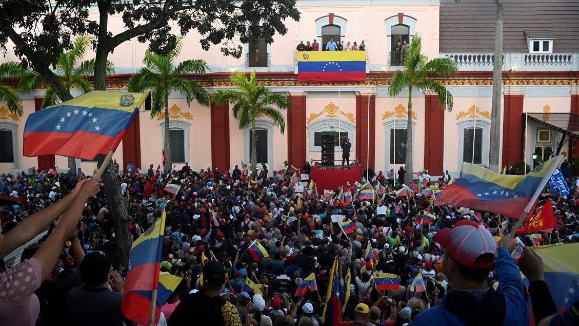 A imagem mostra dezenas de pessoas com bandeiras da Venezuela em frente a um prédio estilo colonial cor de rosa claro. Na sacada central, Maduro fala.