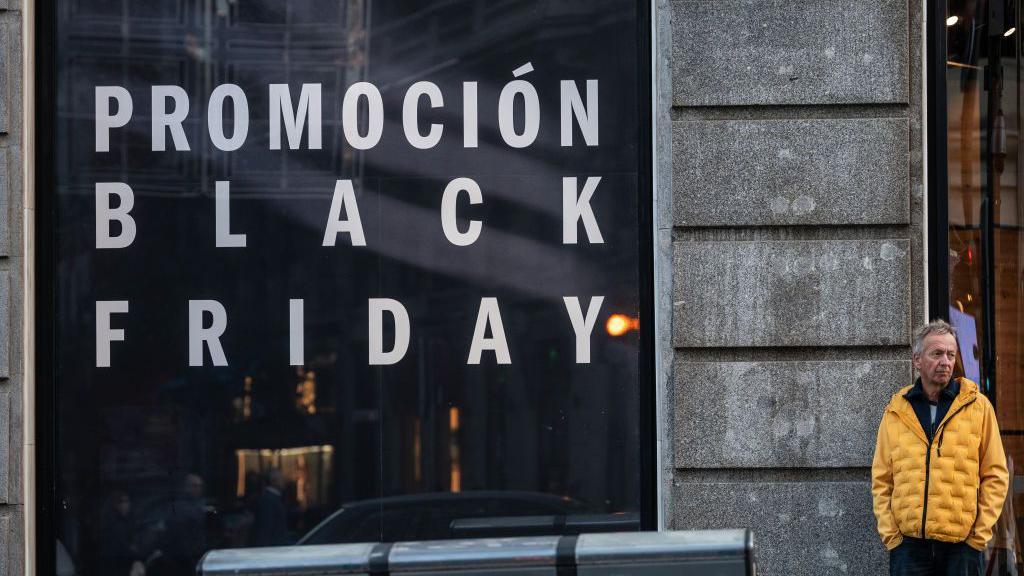 Un hombre con una chaqueta amarilla espera frente a una tienda que ofrece descuentos de Black Friday