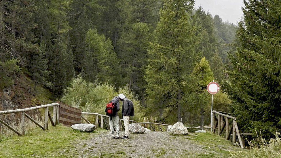 شابان يقفان على مسار جبلي مرصوف بالحصى على الجانب الإيطالي من الحدود الفرنسية الإيطالية، يرتديان حذاء رياضي ويحملان حقيبة صغيرة