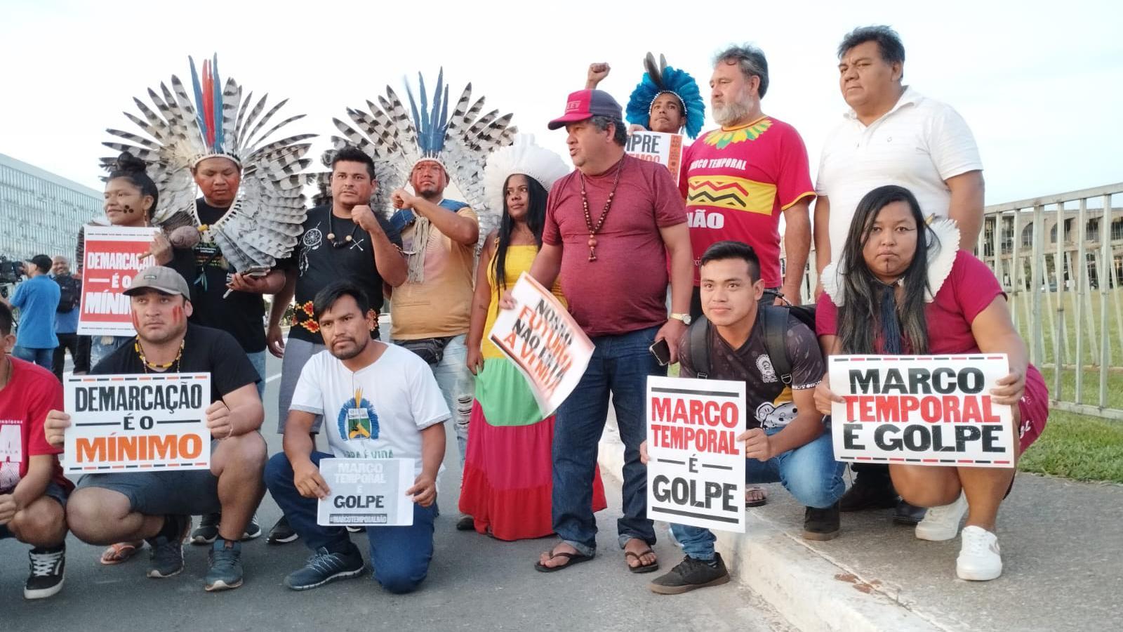 Pessoas segurando cartazes em área externa; alguns usam itens indígenas, como cocares