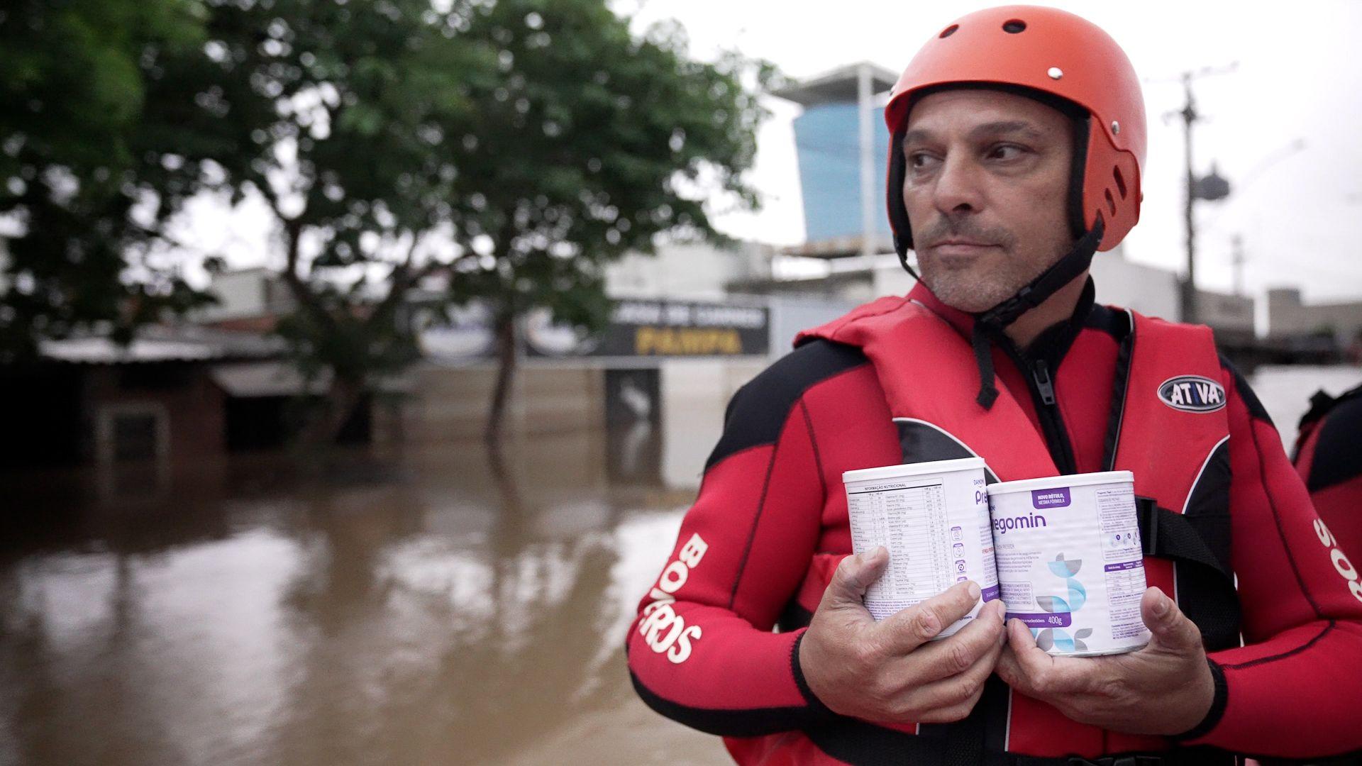 Homem em barco com colete e capacete e latas de leite em pó na mão em uma rua alagada