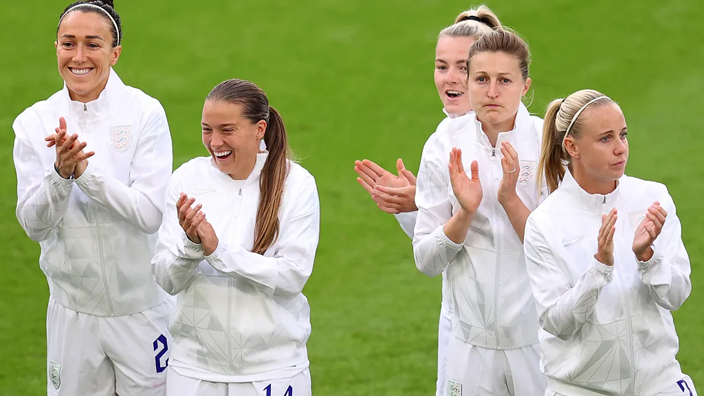 Jogadoras de futebol vestindo uniformes brancos aplaudem enquanto sorriem