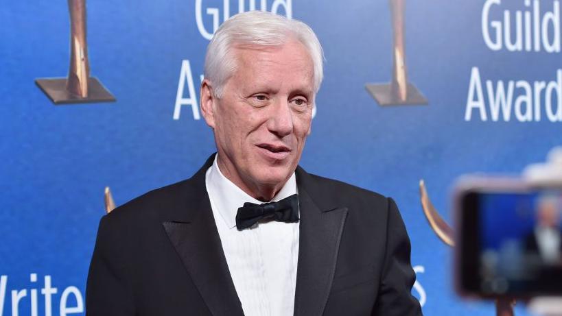 James Woods em traje formal sorrindo em frente ao painel do evento 