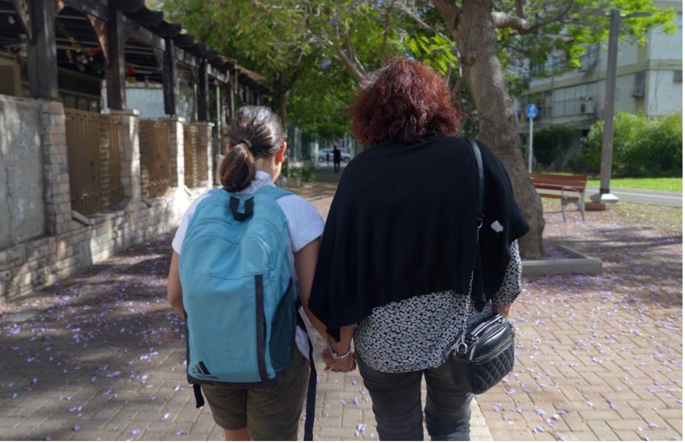 Vista traseira de Osher, de 10 anos, vestindo uma mochila, andando pela rua de mãos dadas com Irit.