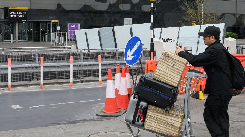 Persona cargada con un trolley lleno de maletas revisa su teléfono a las puertas del aeropuerto de Heathrow, cerrado. 