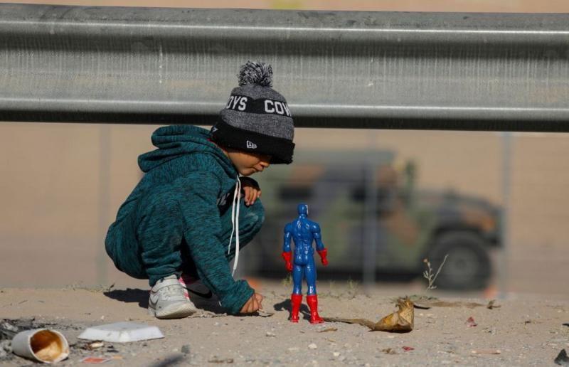 Criança brinca com boneco no chão de areia, com um veículo militar ao longe