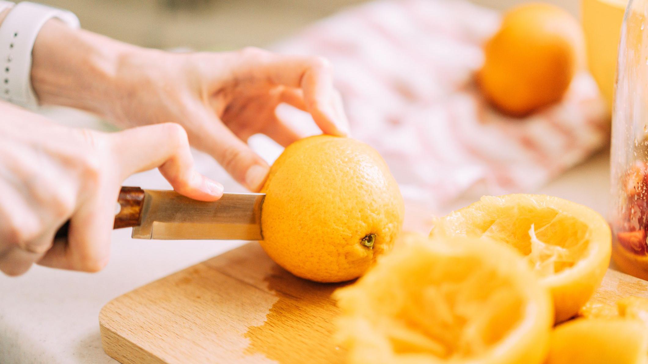 Mãos cortando uma laranja com faca sobre uma tábua de madeira, com laranjas já espremidas ao lado