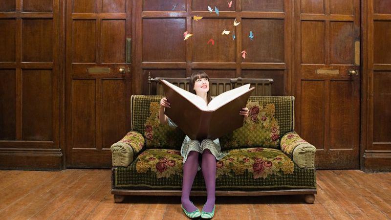 Mulher sentada com livro gigante na mão olhando para o alto e vendo borboletas coloridas