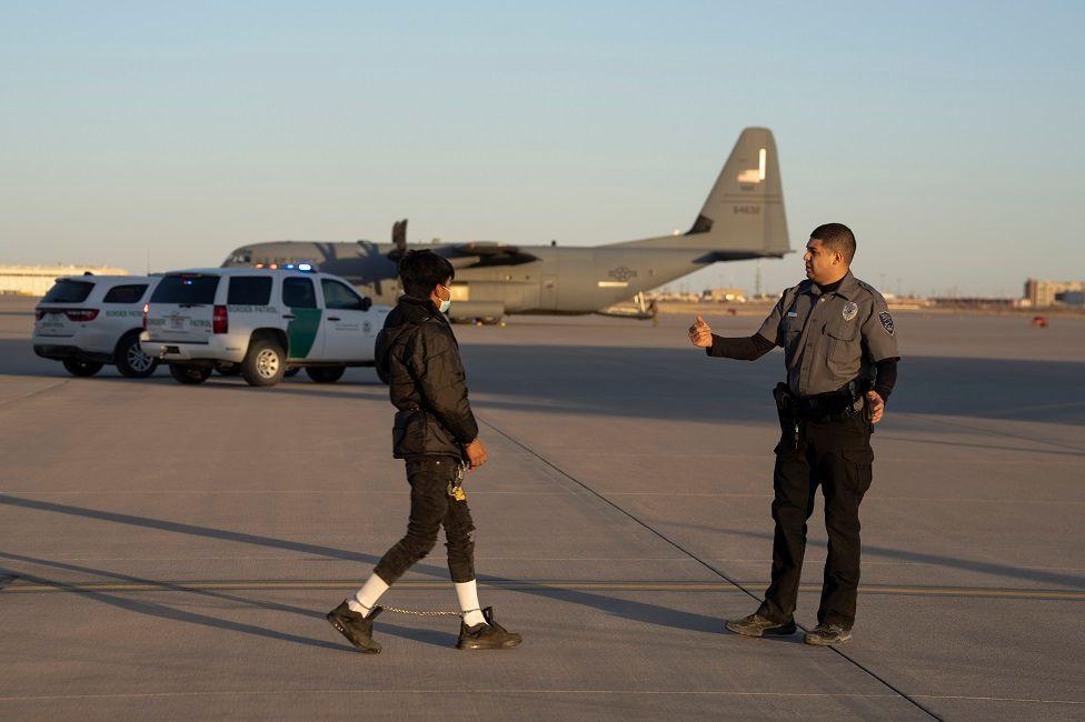 Un joven con esposas y una cadena entre los pies caminando siendo guiado por un funcionario en un aeropuerto