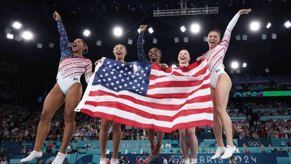 Biles e companheiras festejando o ouro com uma bandeira dos EUA