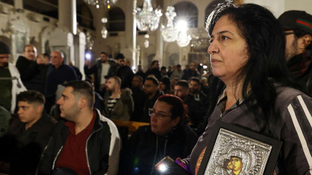 O interior de uma igreja, cheio de gente. Em primeiro plano à direita, uma mulher segura uma placa representando a Virgem Maria e Jesus