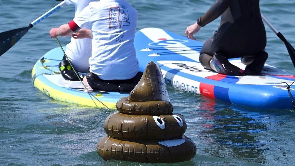 Emoticon de cocô inflável boia na água próximo a pranchas de stand-up paddle 