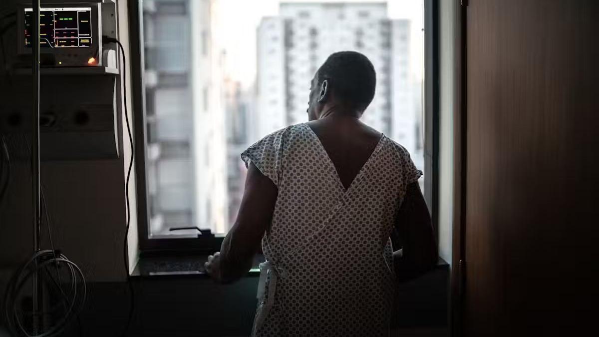 Paciente com avental hospitalar olhando pela janela, mãos apoiadas no parapeito