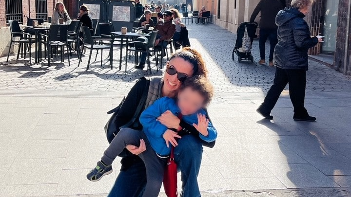 Fotografia colorida mostra mulher branca com uma criança no colo em uma rua de tijolos só para pedestres