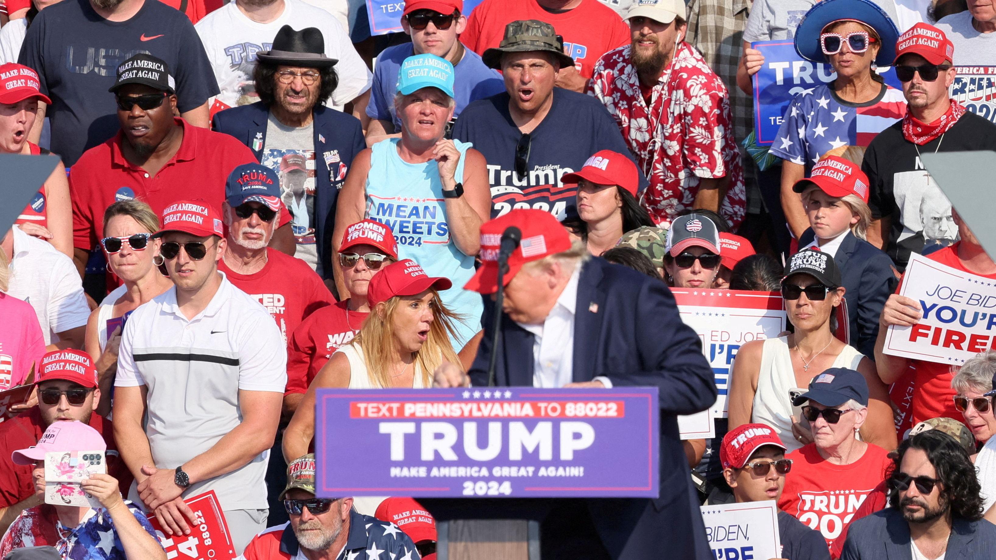 Momento em que as balas são disparadas: Trump está no púlpito, de boné vermelho e com a cabeça abaixada. No fundo, diversos apoiadores com expressão surpresa enquanto seguram cartazes em apoio ao republicano.