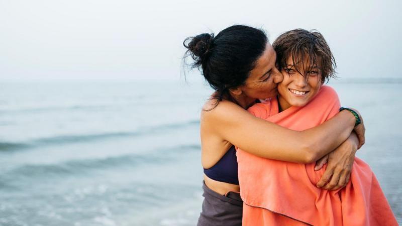 Mulher branca abraça e beija um adolescente branco envolto em uma toalha, no mar, ele sorri