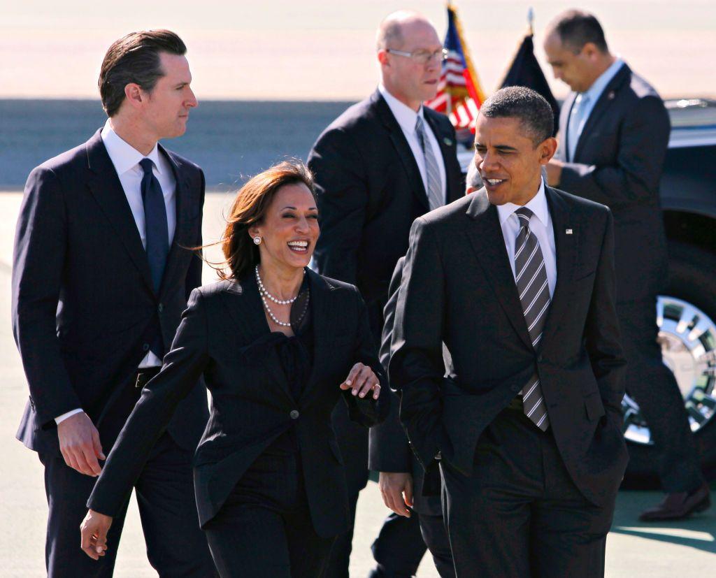 Obama e Harris conversando e sorrindo em estacionamento, acompanhados por assessores e seguranças 