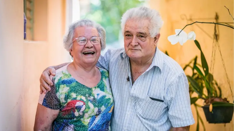 Mulher idosa, cabelo brancos e usando óculos, abraça homem idoso, de cabelos brancos, com camisa