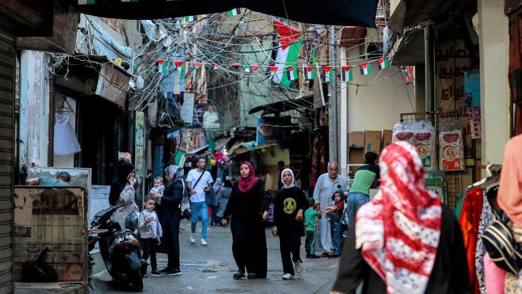 Pedestres caminhando em viela com várias pequenas bandeiras palestinas penduradas