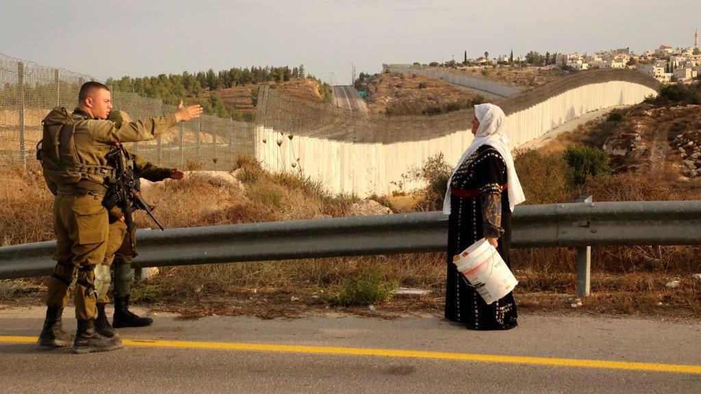معركة حزب الله لا تتعلق بمساعدة الفلسطينيين - في الغارديان 1 فلسطينية تقف أمام جنود إسرائيليين بالقرب من ضواحي مدينة الخليل في الضفة الغربية المحتلة، وفي الخلف يظهر الجدار العازل الإسرائيلي.