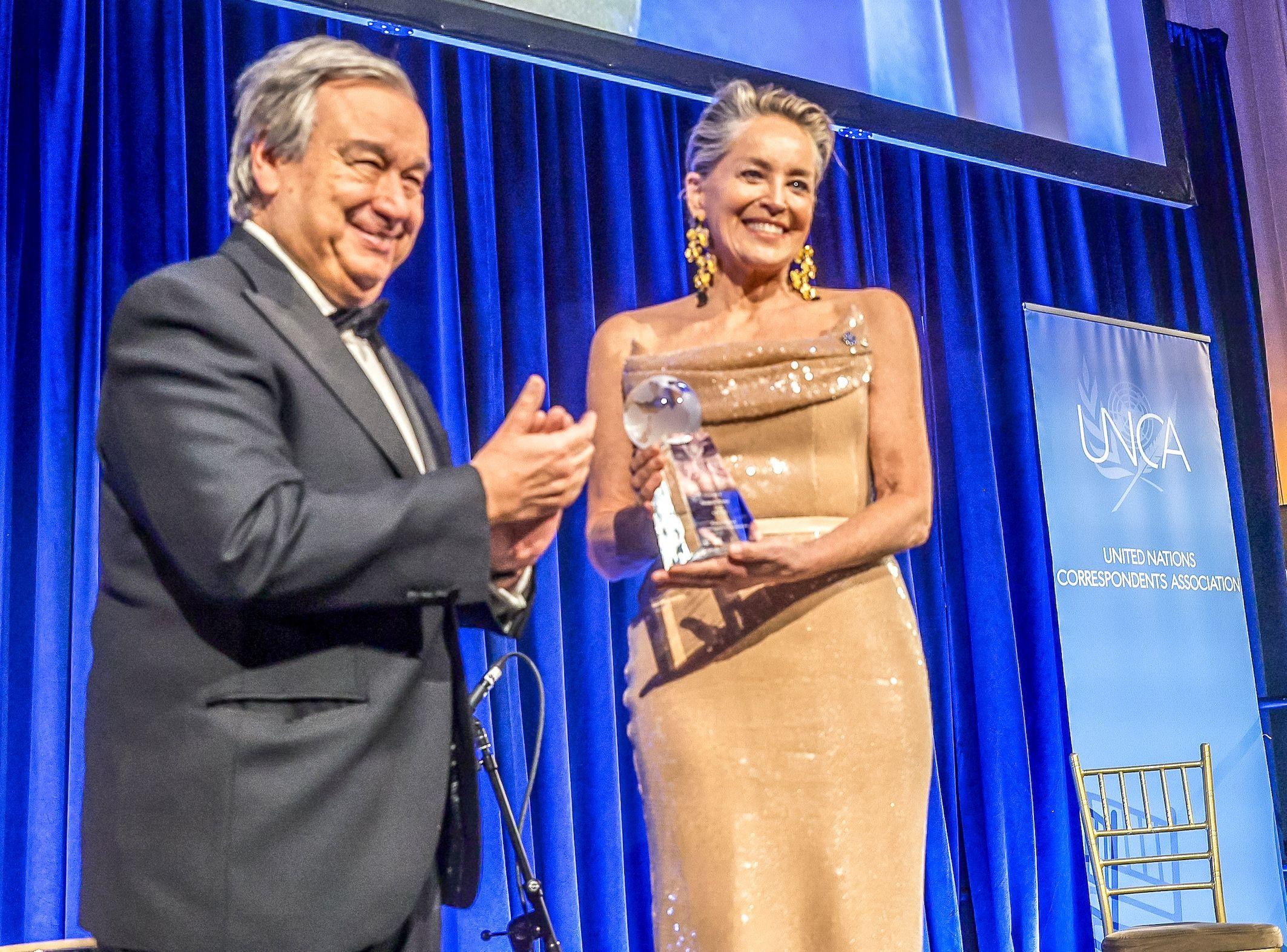 Sharon Stone, con un vestido dorado, sostiene un premio de cristal junto al Secretario General de la ONU, António Guterres, quien sonríe y aplaude.