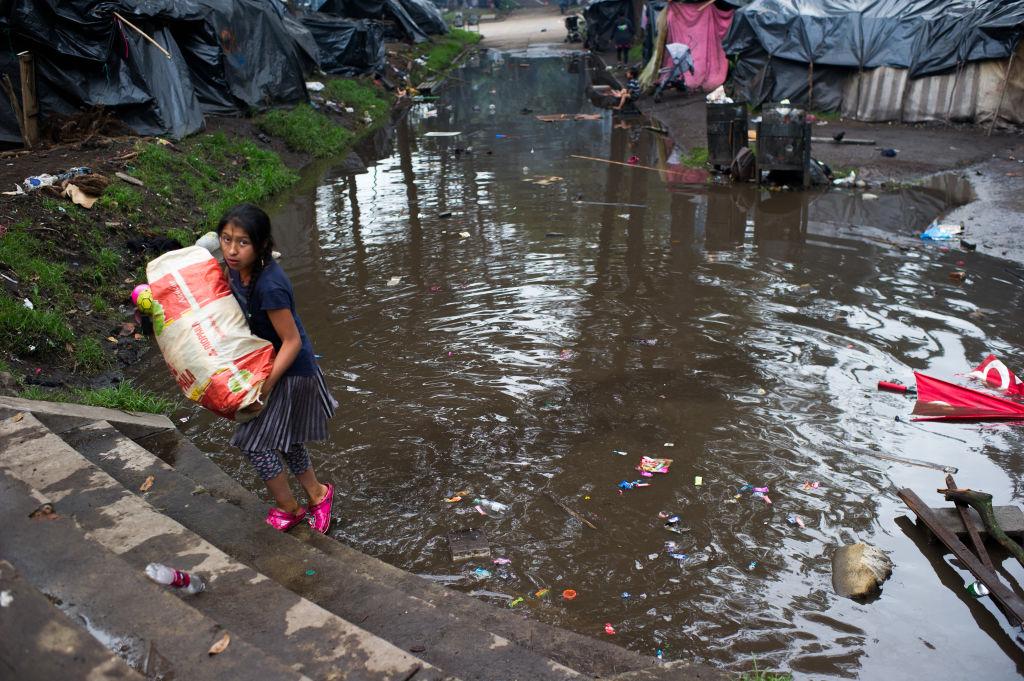 Em Bogotá, na Colômbia, menina indígena atravessa poça carregando uma cesta básica, em acampamento improvisado como abrigo para indígenas deslocados por conflitos (9 de maio de 2022)