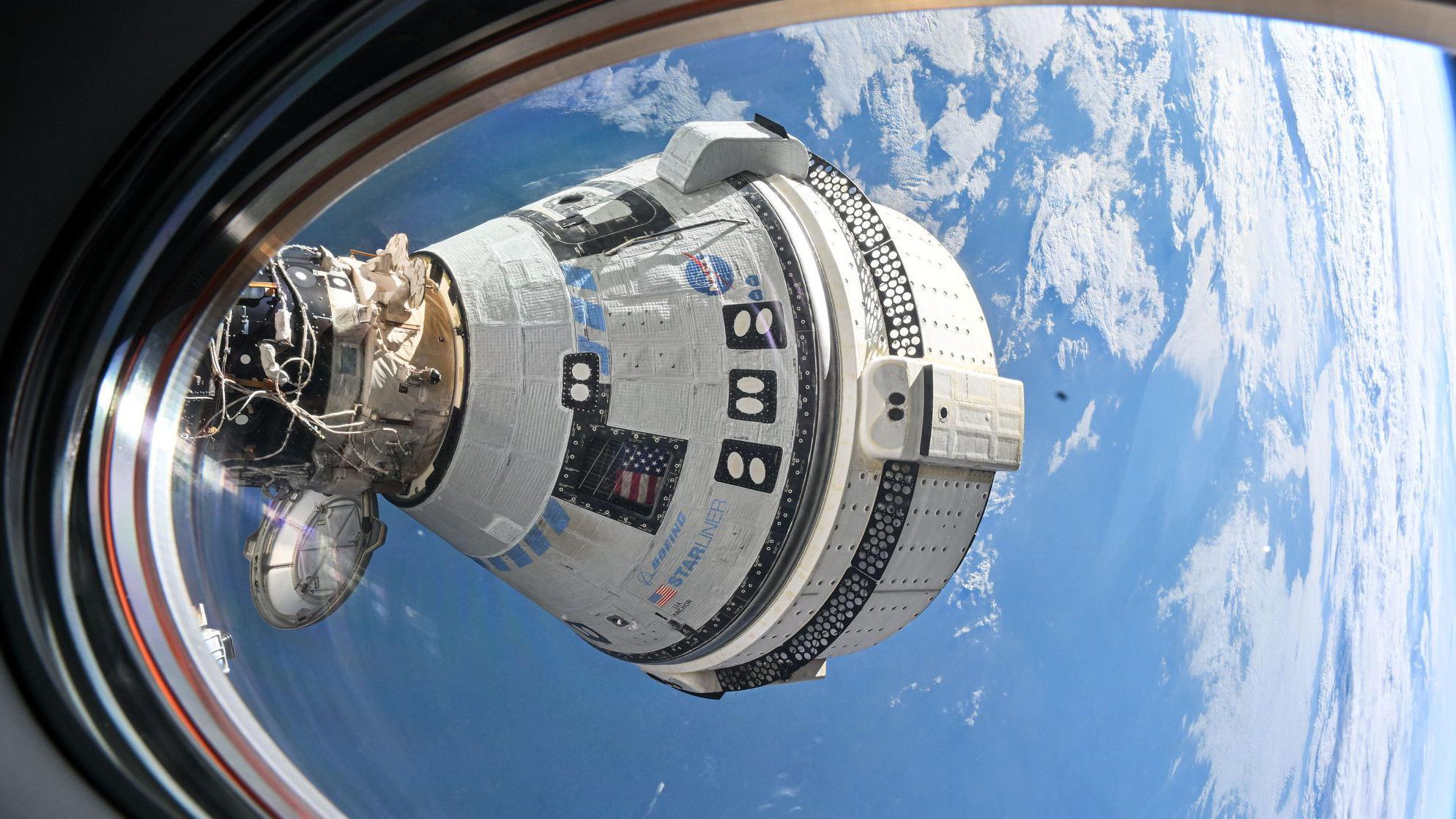 Cápsula Starliner vista da janela da Estação Espacial Internacional - o detalhe branco da nave é claro, com o logo da NASA e a bandeira americana visíveis em alguns lugares. O que parece ser nuvens e a superfície azul da Terra são visíveis ao fundo.