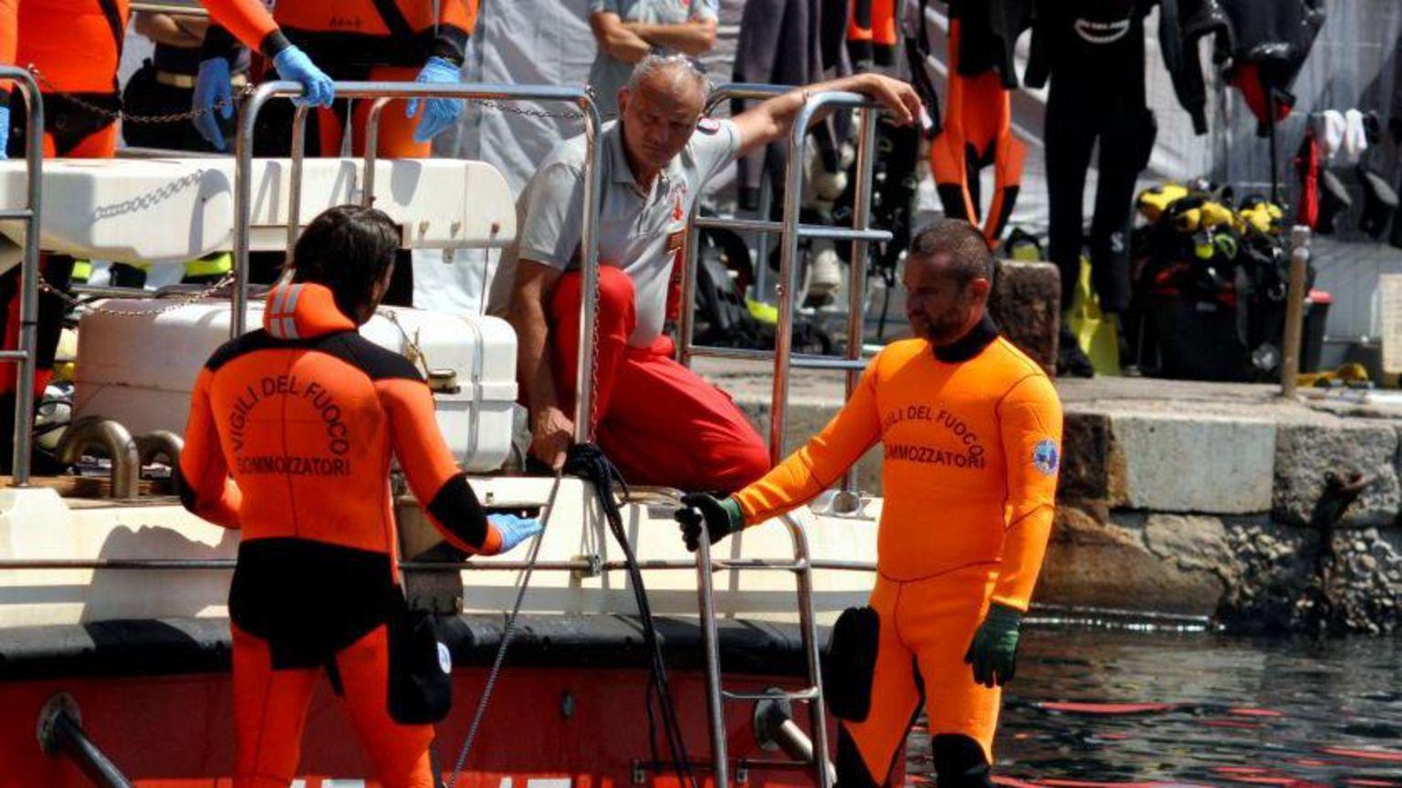 A foto mostra dois homens vestidos com roupas de mergulho laranja na beira de um barco em um porto. 