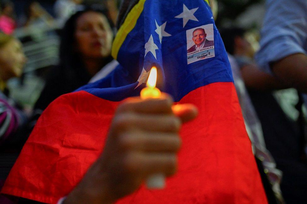 Una persona con una vela encendida y la cara cubierta con la bandera de Venezuela y la foto de Edmundo González