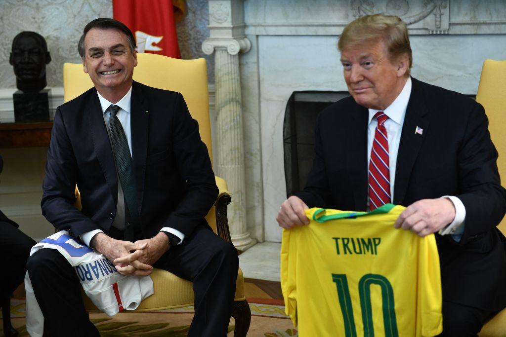 Trump segurando uma camisa do Brasil estampada com o nome dele sentado ao lado de Bolsonaro