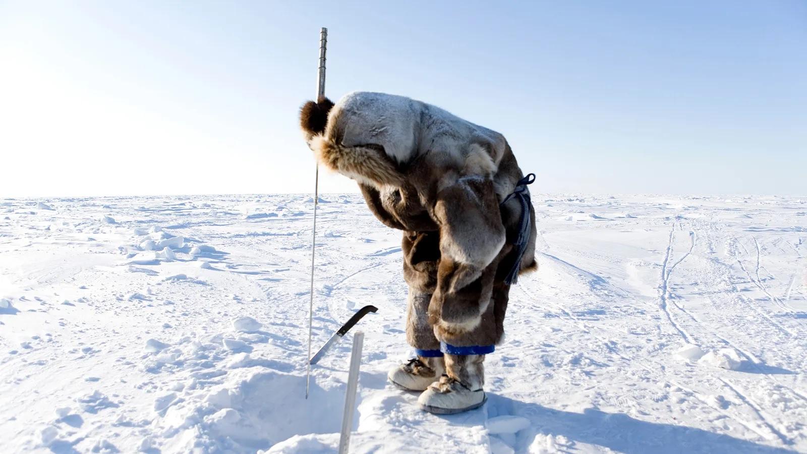 Foto mostra pessoa com roupa de frio pescanso com arpão em um buraco no gelo