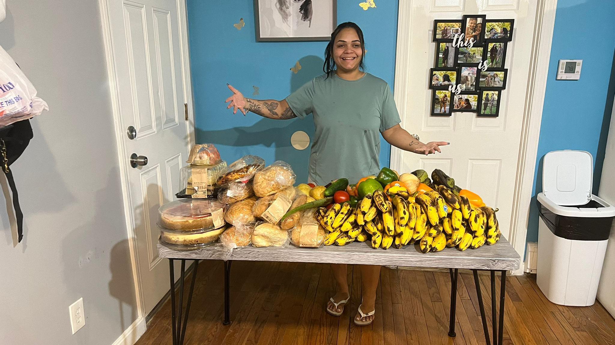 Mesa com pães, bolos e bananas, e uma mulher de pele morena atrás 